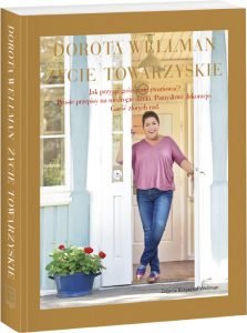 zdjęcie okładki, na której Dorota Wellman stoi w wejściu do swojego domu, ma super kolorowe buty i gustowna bluzką w kolorze różowym